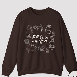 May include: Dark chocolate-colored sweatshirt with a white graphic design of baking tools and the text "I'll be in my office." The design includes a mixer, whisk, apron, and other kitchen items. The sweatshirt has a crew neck and long sleeves.