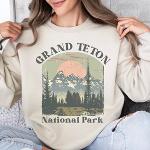 May include: A beige sweatshirt with a vintage-style graphic of the Grand Teton National Park. The graphic features a mountain range with a pink sunset and the text "Grand Teton National Park".