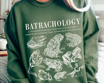 May include: A green sweatshirt with the text "BATRACHOLOGY is the branch of zoology concerned with the study of amphibians including frogs, salamanders, and caecilians" printed in white. The sweatshirt also features a white illustration of various frogs and toads. The text "MILITARY GREEN" is printed in white at the bottom right corner of the sweatshirt.