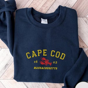 May include: Navy blue sweatshirt with embroidered text "CAPE COD" in yellow, a red lobster graphic, and the text "16 02 MASSACHUSETTS". The sweatshirt has a crew neck and long sleeves. The color is navy.