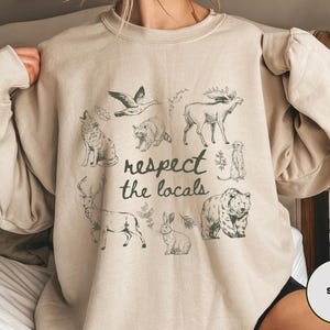 May include: Sand-colored sweatshirt with a graphic of various animals including a wolf, moose, and bear. The text "respect the locals" is printed in the center. The sweatshirt is a neutral color.