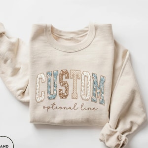 May include: A sand-coloured sweatshirt featuring the word "CUSTOM" in a collegiate font. The letters are a mix of floral and patterned designs. The text "optional line" is embroidered below the word "CUSTOM".
