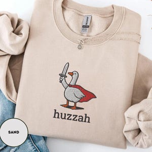 May include: Sand-coloured sweatshirt with an embroidered cartoon goose wearing a red cape and holding a sword. The word "huzzah" is printed below the goose. A silver necklace is draped on the sweatshirt.