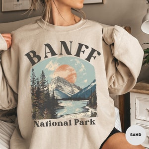 May include: A beige sweatshirt with a vintage-style graphic of a mountain range, a lake, and a sun. The graphic is surrounded by a black circle with the text "BANFF National Park" in black lettering.