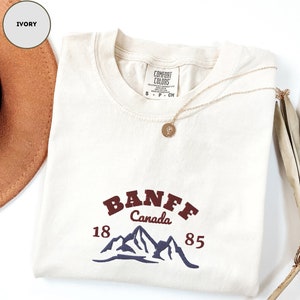 May include: Ivory t-shirt with a brown and blue graphic of mountains and the text "Banff Canada 18 85". The shirt has a tag that says "Comfort Colors".