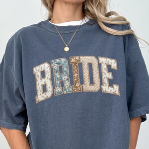 May include: A denim blue oversized t-shirt with the word "BRIDE" in a faux patchwork design across the chest. The design incorporates floral and tan patterns. The shirt has short sleeves and a relaxed fit. The text "PLEASE NOTE: THIS IS A DTG-PRINTED FAUX PATCHWORK DESIGN, NOT REAL STITCHED OR SEWN PATCHES." is at the bottom.