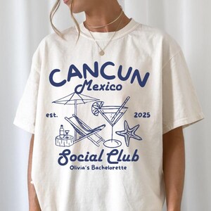 May include: Ivory t-shirt with a blue graphic design that says "Cancun Mexico est. 2025 Social Club Olivia's Bachelorette" with a cocktail glass, beach chair, and starfish.