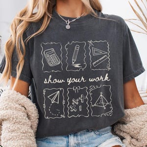 May include: Dark gray t-shirt with white line drawings of a calculator, pencil, ruler, paper airplane, math symbols, and a pyramid. The text "show your work" is below the drawings. The shirt is worn with denim shorts and a cream-colored cardigan.