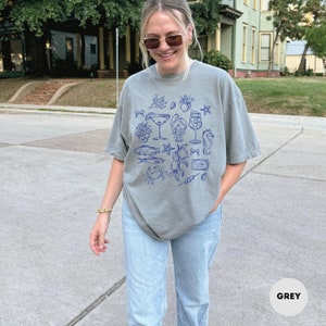 May include: A grey t-shirt with a blue illustration of seafood and cocktails. The design includes images of fish, a lobster, a seahorse, and various cocktail glasses. The word "GREY" is printed in a white box at the bottom.