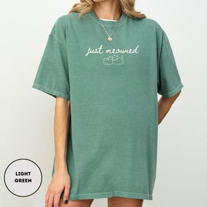 May include: Light green t-shirt with the text "just meowied" in a cursive font, along with a small illustration of two cats. The shirt is a relaxed fit and has short sleeves.