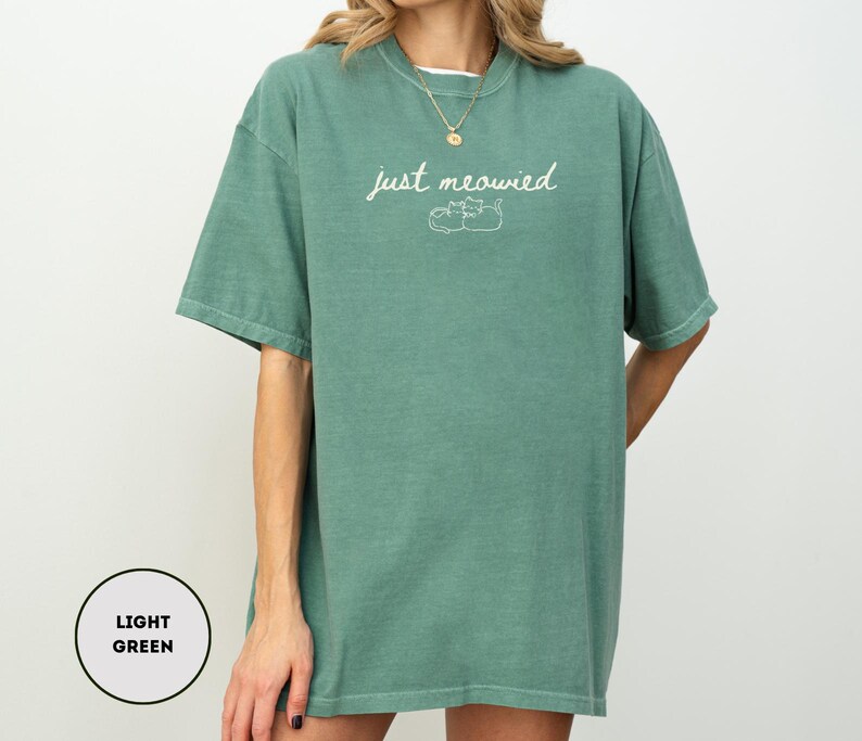 May include: Light green t-shirt with the text "just meowied" in a cursive font, along with a small illustration of two cats. The shirt is a relaxed fit and has short sleeves.