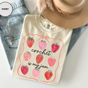 Puede incluir: Camiseta de color marfil con un dise&ntilde;o de fresas y el texto "crochet is my jam". La camiseta est&aacute; colocada sobre unos vaqueros azul claro. Tambi&eacute;n se ve un sombrero beige y un collar.
