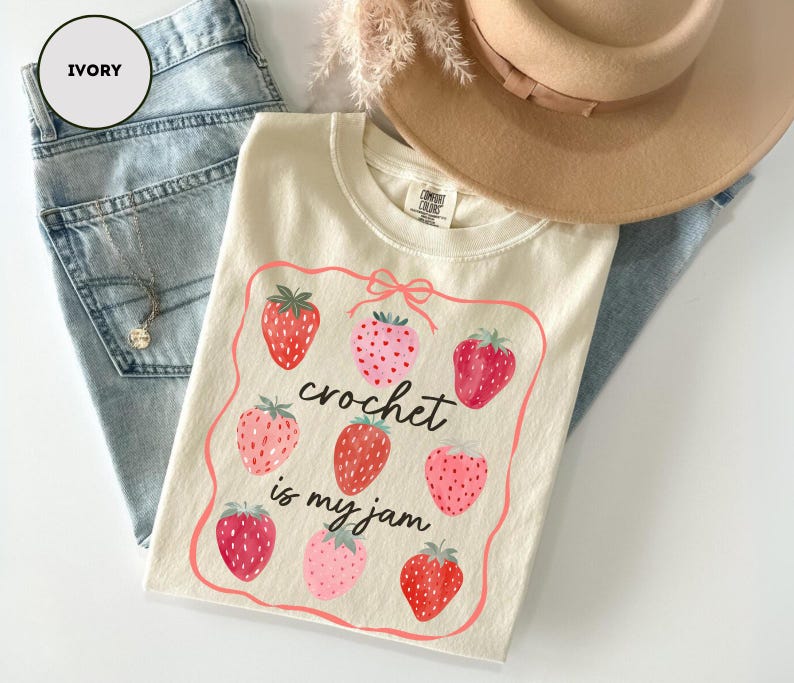 Puede incluir: Camiseta de color marfil con un dise&ntilde;o de fresas y el texto "crochet is my jam". La camiseta est&aacute; colocada sobre unos vaqueros azul claro. Tambi&eacute;n se ve un sombrero beige y un collar.