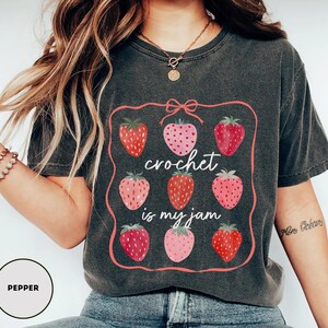 Puede incluir: Camiseta gris oscuro con un dise&ntilde;o enmarcado en rosa con nueve fresas. El texto "crochet is my jam" est&aacute; en el centro. Las fresas son en tonos rojos y rosas. La camiseta es de estilo casual.
