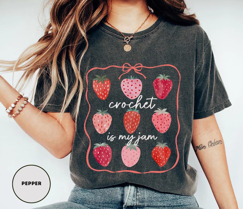 Puede incluir: Camiseta gris oscuro con un dise&ntilde;o enmarcado en rosa con nueve fresas. El texto "crochet is my jam" est&aacute; en el centro. Las fresas son en tonos rojos y rosas. La camiseta es de estilo casual.