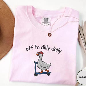May include: A light pink Comfort Colors t-shirt with the embroidered phrase "off to dilly dally" above a goose riding a scooter. The goose wears a red cap. A gold necklace and a brown hat are also visible.