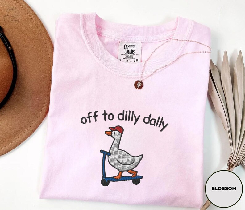 May include: A light pink Comfort Colors t-shirt with the embroidered phrase "off to dilly dally" above a goose riding a scooter. The goose wears a red cap. A gold necklace and a brown hat are also visible.
