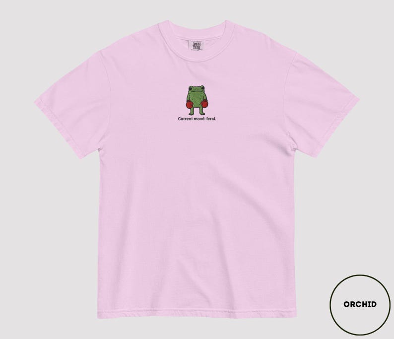 May include: A light pink t-shirt featuring a green frog wearing red boxing gloves. The text "Current mood: feral" is below the frog. The shirt is a comfortable, casual style.