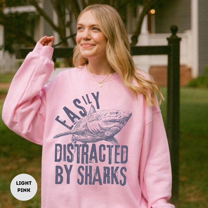 May include: A light pink sweatshirt with a graphic of a shark and the text "Easily Distracted By Sharks".