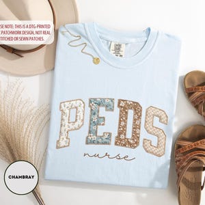 May include: Light blue t-shirt with "PEDS nurse" text. The word "PEDS" is in a patchwork design with floral and textured patterns. A gold necklace and brown sandals are also in the image. A hat and decorative plant are in the background.