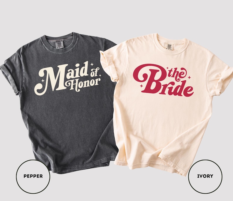 May include: Two t-shirts, one in pepper and one in ivory. The pepper-coloured shirt says "Maid of Honor" in a retro font. The ivory shirt says "The Bride" in a similar font. Both are short-sleeved.