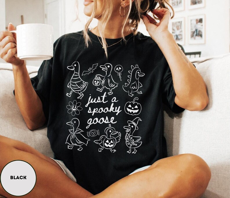 May include: Black t-shirt with a white graphic of cartoon geese in Halloween costumes and the text "just a spooky goose". The design includes a pumpkin, bats, and candy. The shirt is a comfortable, casual style.