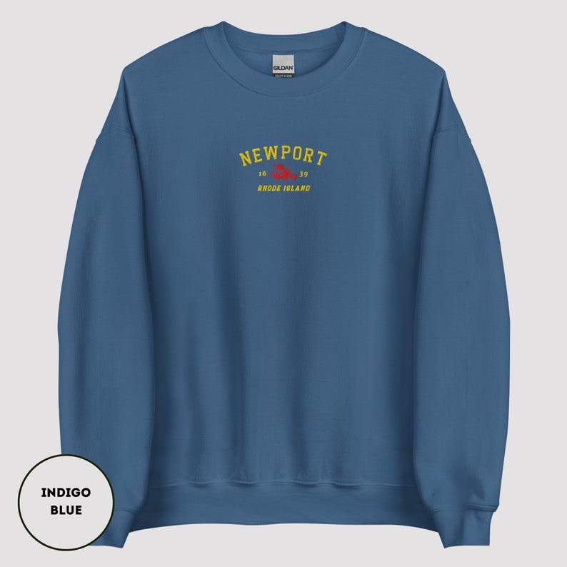 May include: Indigo blue sweatshirt featuring the word "NEWPORT" in yellow arched text, with "16 39" and "RHODE ISLAND" below. The sweatshirt has a crew neck and long sleeves. The lower left corner has a circle with the words "INDIGO BLUE".