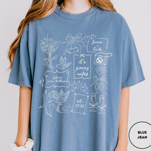 May include: A blue jean-colored t-shirt with white line art. The design includes flowers, bows, doves, a camera, champagne glasses, and the text "it's giving wifey", "future mrs. Williams", "James + Emily", and "est. 2025".