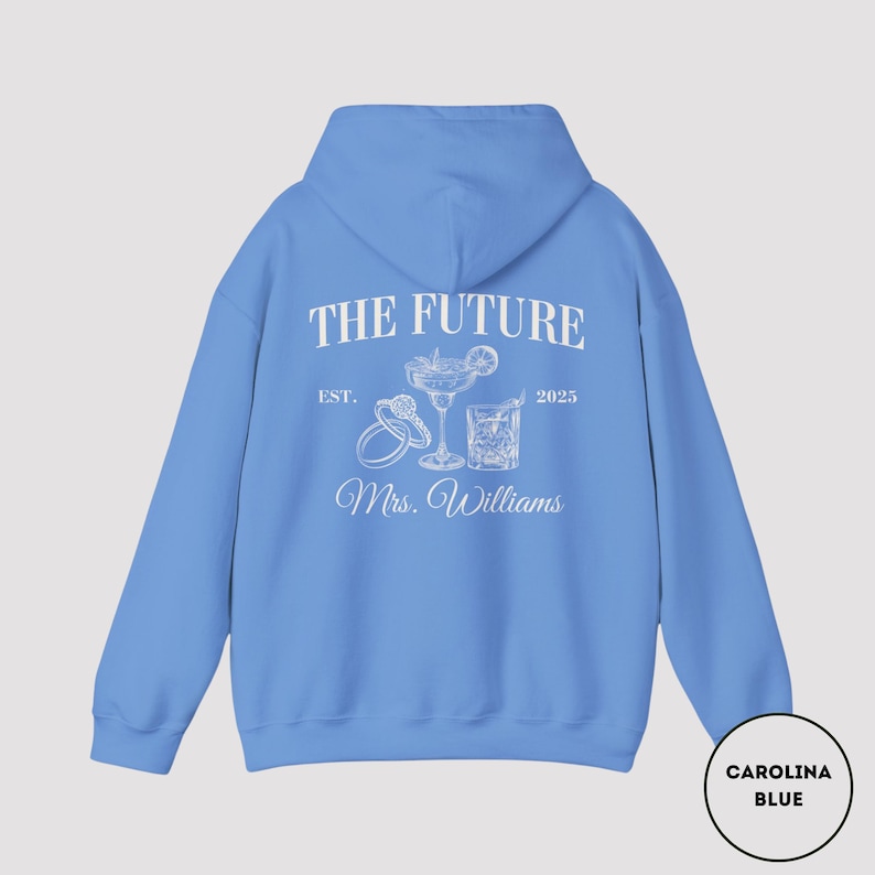 Pode incluir: Camisola de capuz azul claro com texto branco que diz "The Future Est. 2025 Mrs. Williams" com um desenho de dois copos de cocktail e um anel.