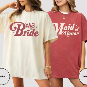May include: Two oversized t-shirts, one ivory and one crimson. The ivory shirt has the text "the Bride" in a retro font. The crimson shirt says "Maid of Honor". Both shirts are short-sleeved and suitable for a wedding.