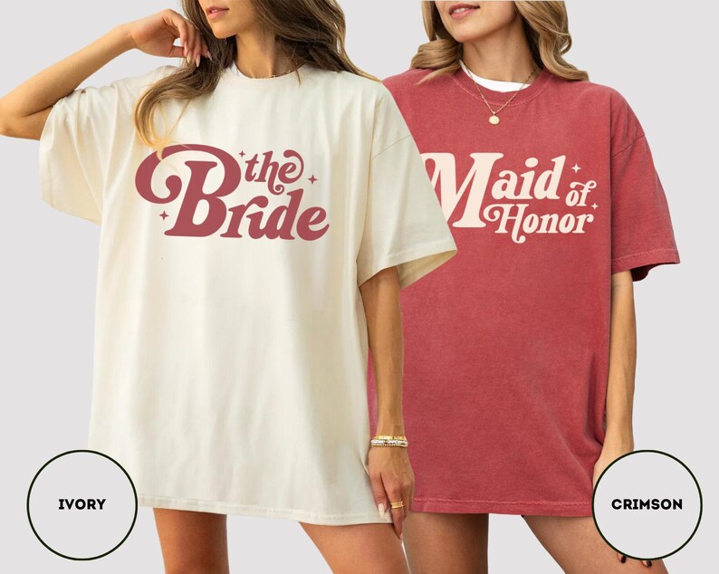 May include: Two oversized t-shirts, one ivory and one crimson. The ivory shirt has the text "the Bride" in a retro font. The crimson shirt says "Maid of Honor". Both shirts are short-sleeved and suitable for a wedding.