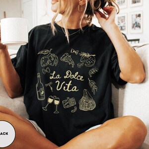 May include: Black t-shirt with a gold graphic design featuring the words "La Dolce Vita" and illustrations of food and wine. The design includes a bottle, wine glasses, and various food items. The shirt is a relaxed fit.