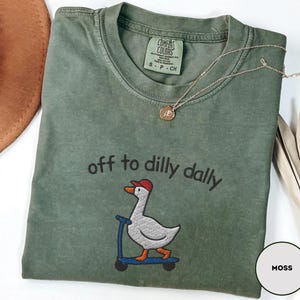 May include: Moss green t-shirt with the embroidered phrase "off to dilly dally" and a cartoon goose on a scooter. The shirt has a crew neck and short sleeves. A gold necklace with a pendant is draped on the shirt.