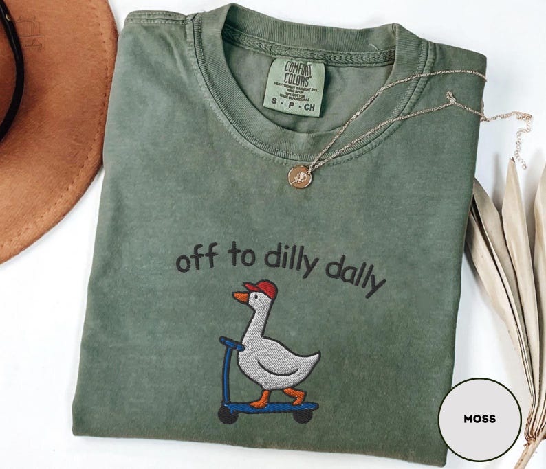 May include: Moss green t-shirt with the embroidered phrase "off to dilly dally" and a cartoon goose on a scooter. The shirt has a crew neck and short sleeves. A gold necklace with a pendant is draped on the shirt.