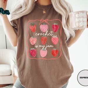 Puede incluir: Camiseta color espresso con un dise&ntilde;o de fresas y el texto "crochet is my jam". El dise&ntilde;o incluye nueve fresas en varios tonos de rojo y rosa, enmarcadas por un borde rosa. La camiseta es de estilo c&oacute;modo e informal.