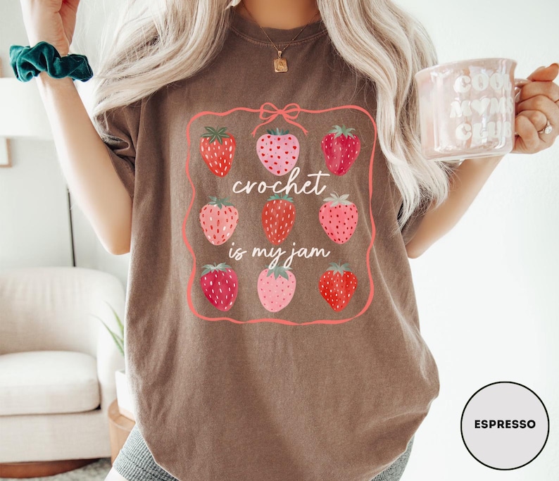 Puede incluir: Camiseta color espresso con un dise&ntilde;o de fresas y el texto "crochet is my jam". El dise&ntilde;o incluye nueve fresas en varios tonos de rojo y rosa, enmarcadas por un borde rosa. La camiseta es de estilo c&oacute;modo e informal.