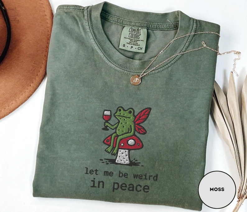 May include: Moss green t-shirt with a whimsical embroidered design. The design features a green frog with fairy wings, holding a wine glass, seated on a red mushroom. The text "let me be weird in peace" is below the frog. A gold necklace is draped on the shirt.