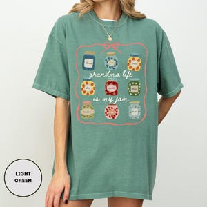Peut inclure: T-shirt vert clair avec un motif de bocaux de confiture et le texte "grandma life is my jam" dans un cadre rose. Les bocaux pr&eacute;sentent diff&eacute;rents motifs et couleurs, ajoutant une touche ludique au design.