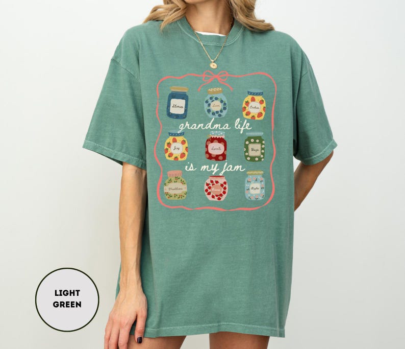 Peut inclure: T-shirt vert clair avec un motif de bocaux de confiture et le texte "grandma life is my jam" dans un cadre rose. Les bocaux pr&eacute;sentent diff&eacute;rents motifs et couleurs, ajoutant une touche ludique au design.