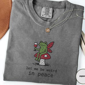 May include: Pepper-colored t-shirt with an embroidered design of a green frog with red fairy wings, holding a glass of wine, sitting on a red mushroom. The text "let me be weird in peace" is below the design.