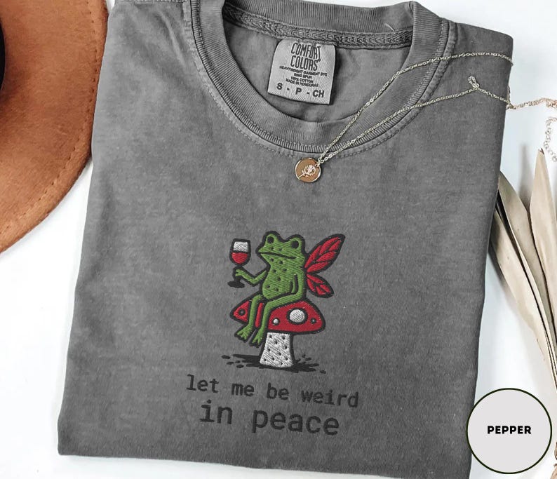 May include: Pepper-colored t-shirt with an embroidered design of a green frog with red fairy wings, holding a glass of wine, sitting on a red mushroom. The text "let me be weird in peace" is below the design.