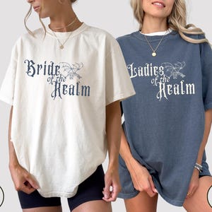 May include: Two short-sleeved t-shirts, one cream and one denim blue, featuring the text "Bride of the Realm" and "Ladies of the Realm" in a gothic font. Each shirt has a matching illustration of a dragon.