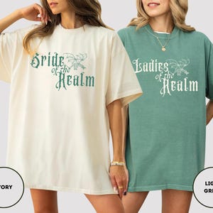 May include: Two oversized t-shirts, one ivory and one light green, each with a crew neck and short sleeves. The ivory shirt features the text "Bride of the Realm" in teal script, while the green shirt says "Ladies of the Realm" in white script.
