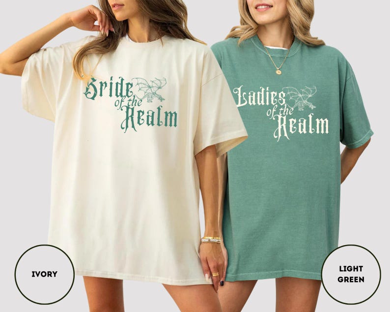 May include: Two oversized t-shirts, one ivory and one light green, each with a crew neck and short sleeves. The ivory shirt features the text "Bride of the Realm" in teal script, while the green shirt says "Ladies of the Realm" in white script.