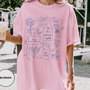 May include: A light pink t-shirt with a blue graphic design featuring bows, doves, and the text "It's giving wifey." Additional text includes "future mrs. Williams," "James & Emily," and "est. 2025." The shirt has a relaxed fit.