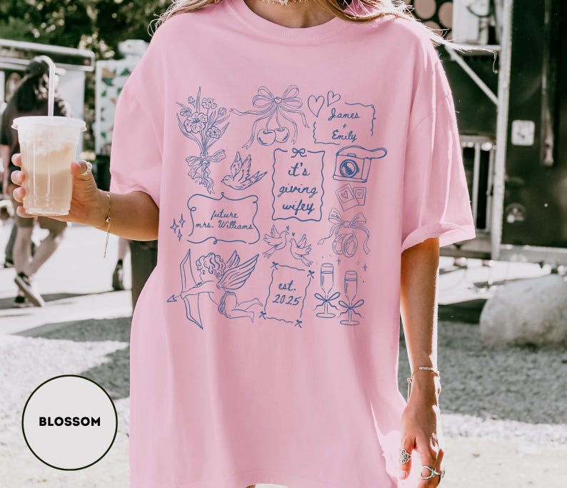 May include: A light pink t-shirt with a blue graphic design featuring bows, doves, and the text "It's giving wifey." Additional text includes "future mrs. Williams," "James & Emily," and "est. 2025." The shirt has a relaxed fit.