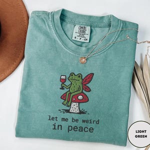 May include: A light green t-shirt with a whimsical frog illustration holding a wine glass, sitting on a mushroom. The text "let me be weird in peace" is printed below the frog. A gold necklace is draped across the shirt.