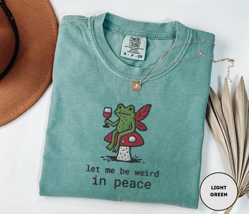 May include: A light green t-shirt with a whimsical frog illustration holding a wine glass, sitting on a mushroom. The text "let me be weird in peace" is printed below the frog. A gold necklace is draped across the shirt.