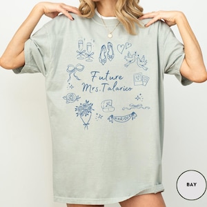 May include: A light green t-shirt with a white collar featuring a blue graphic design. The design includes illustrations of champagne glasses, shoes, hearts, doves, and the text "Future Mrs. Talarico". A bouquet of flowers, a ring, and a banner that says "Bride" are also included.