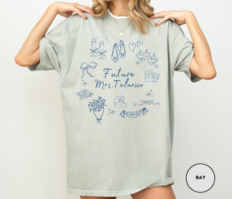 May include: A light green t-shirt with a white collar featuring a blue graphic design. The design includes illustrations of champagne glasses, shoes, hearts, doves, and the text "Future Mrs. Talarico". A bouquet of flowers, a ring, and a banner that says "Bride" are also included.